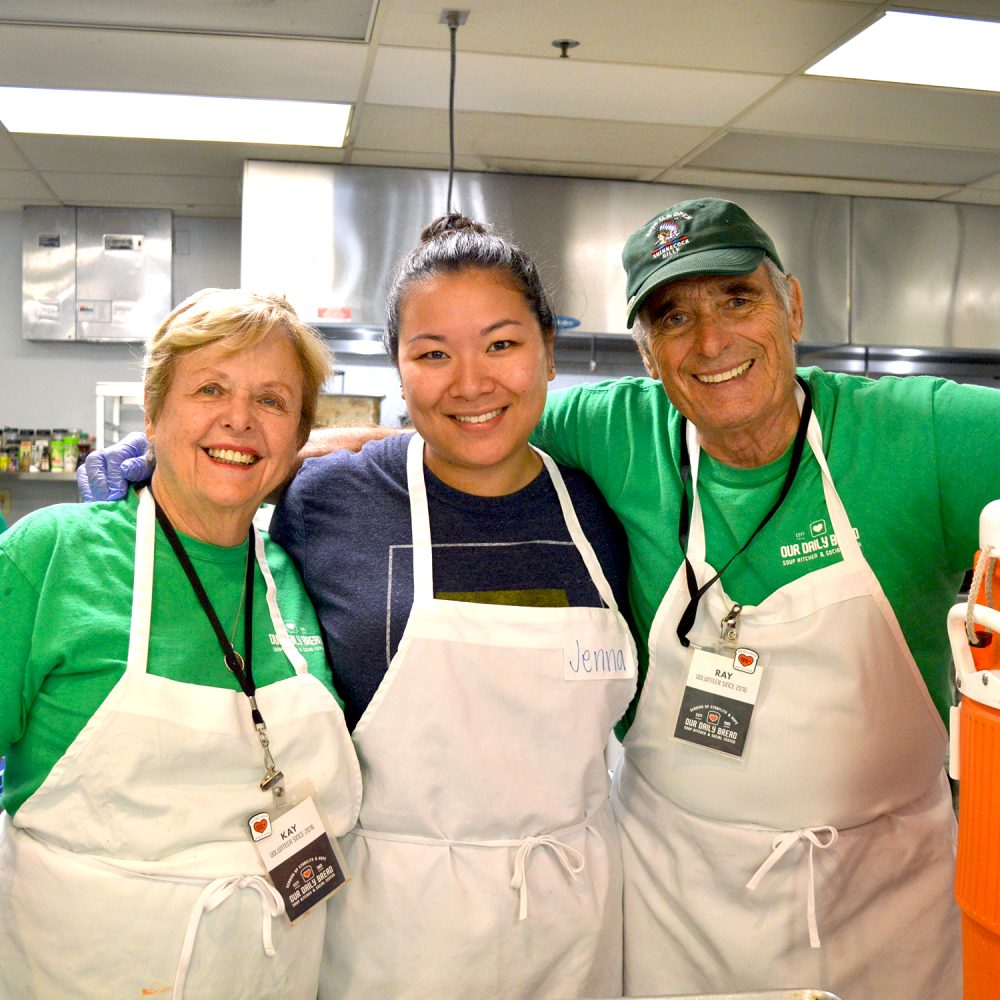 three volunteers standing with arms around each other's shoulders with the kitchen in the background. they are all wearing aprons.