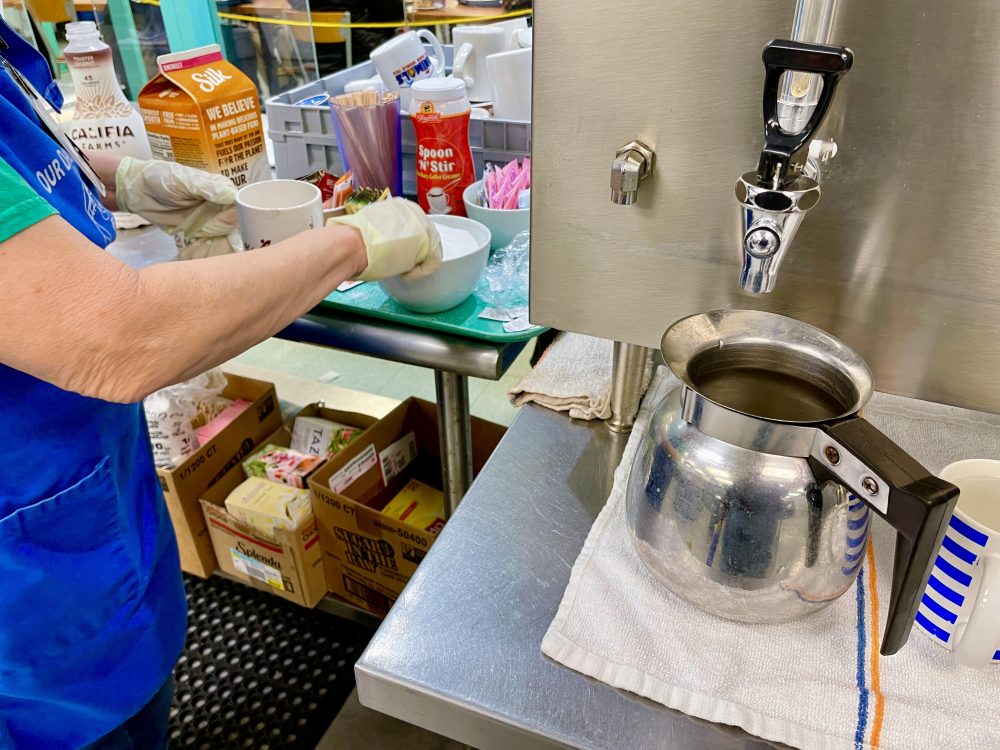 a view of our coffee making station. There is a metal pitcher under a spout, and volunteer arms and hands are showing spooning sugar into a coffee cup with creamer in the background.