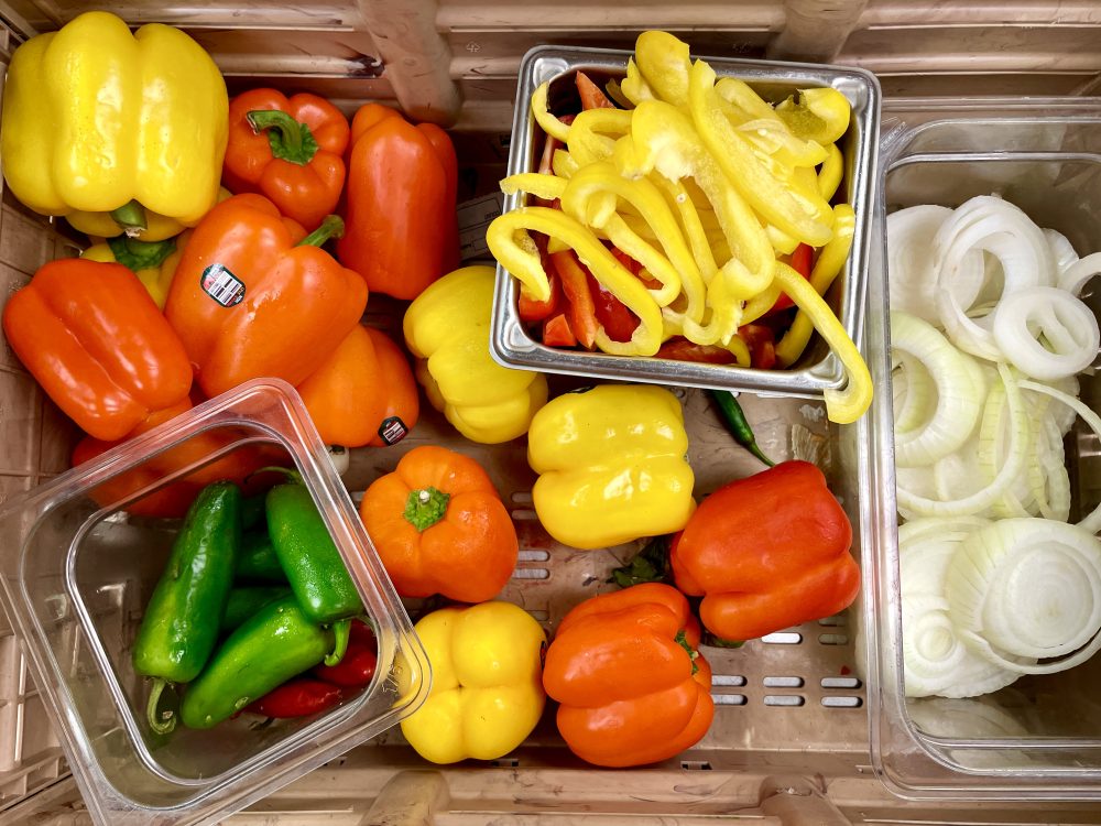 brightly colored bell peppers, spicy peppers, and onions, in various stages of being chopped, all in a wooden bin photographed from above.