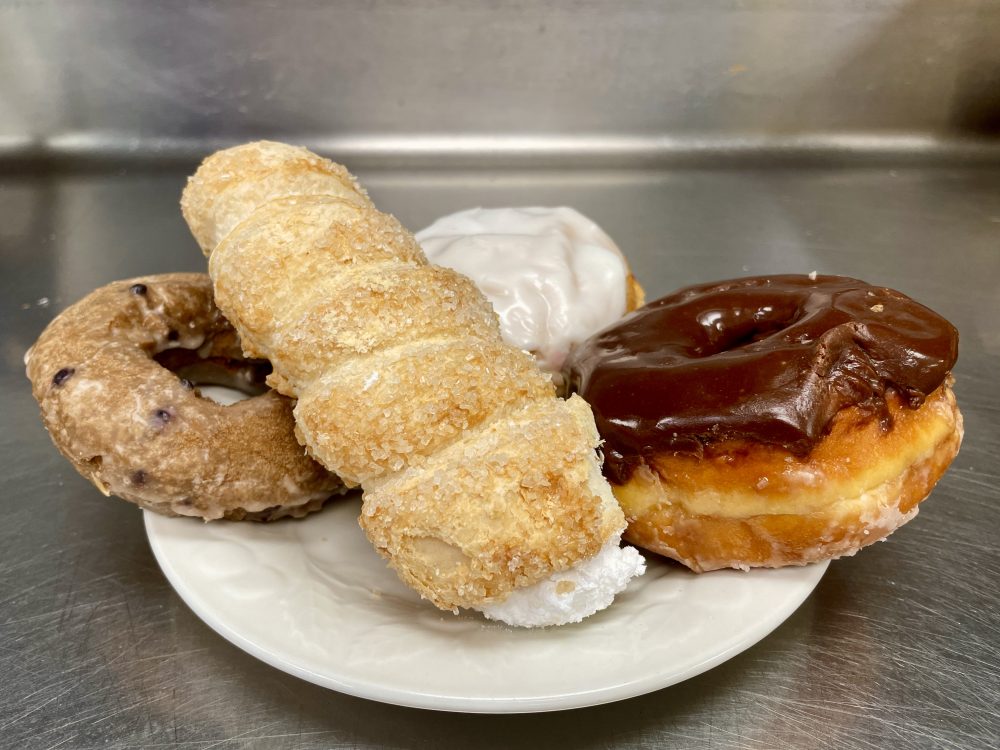 A small white plate sitting on a metal prep counter with four donuts on top. There is a chocolate glazed, one that looks like a cannoli combined with a churro with cream coming out covered in sugar, a glazed blueberry cake one, and peeking out of the back there is one with white icing.
