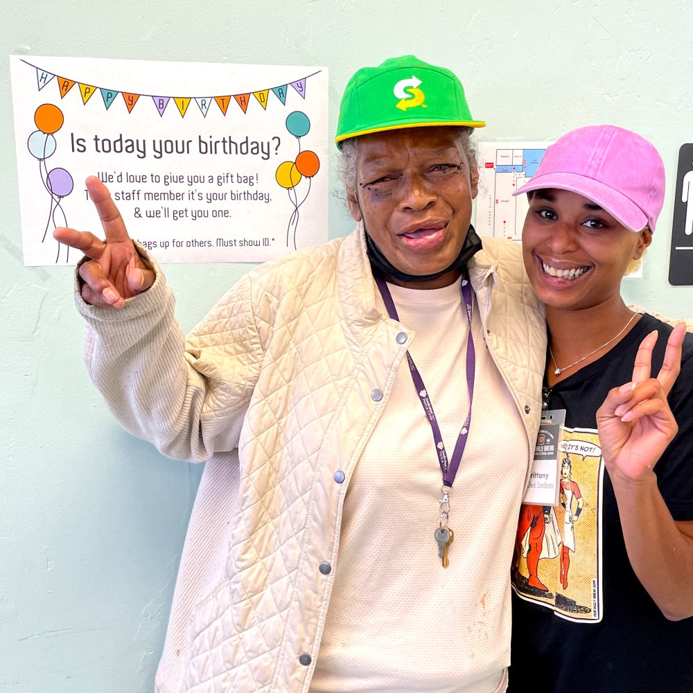 Two women standing side to side with arms around each other, holding up peace signs with their hands and standing next to a sign that says "is today your birthday?" with balloons