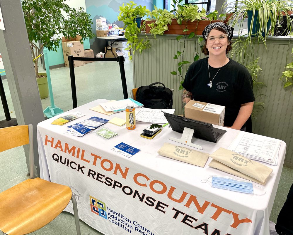 a table in our lower dining room with a tablecloth reading hamilton county quick response team, and information and harm reduction supplies sitting on top. There is a woman sitting on the other side smiling at the camera.