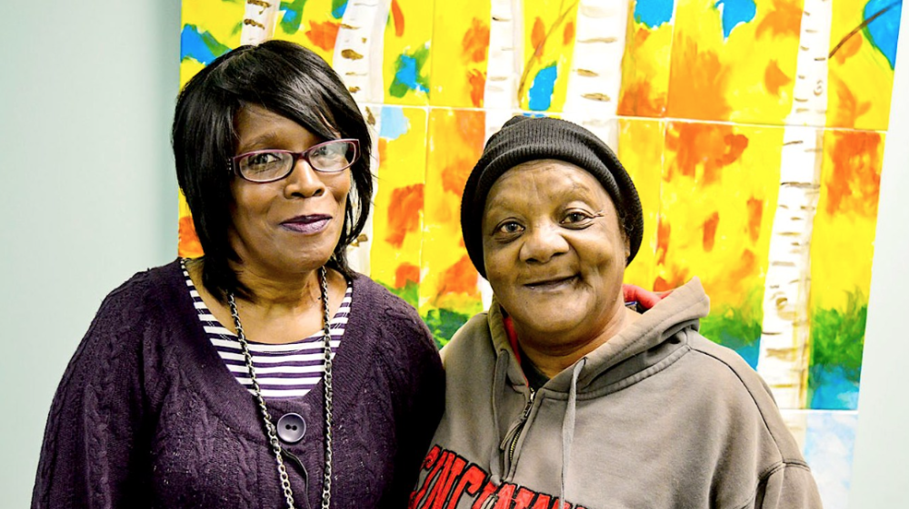 two women standing together smiling at the camera with a landscape painting in the background