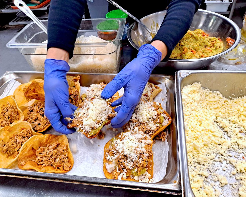 a pair of hands wearing latex gloves are preparing hard shell tacos with cheese, guacamole and meat