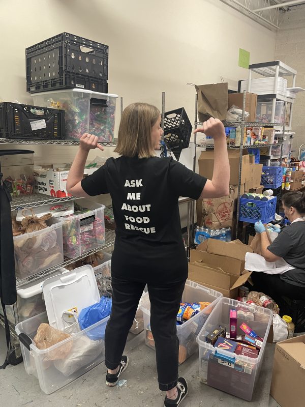 Georgine modeling the t shirt with her back to the camera, thumbs pointing behind her at the back of the tee, which has text that reads ask me about food rescue. she is in our huge pantry surrounded by bins of food and a volunteer sorting.